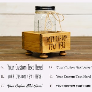 May include: A clear glass jar with a black lid and twine tied around it, set in a small, stained wooden box. The box has the engraved text "YOUR CUSTOM TEXT HERE" on the front and sits on four small, rounded feet. Various text options are listed below.