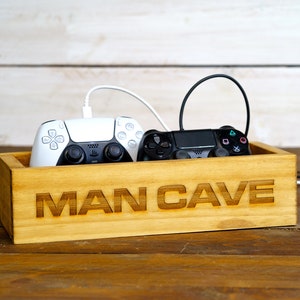 May include: A wooden box with the words "MAN CAVE" engraved on the front. Two video game controllers, one black and one white, are inside the box.
