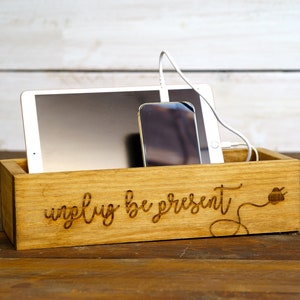 May include: Wooden charging station with the text "unplug be present" engraved on the front. A tablet and a smartphone are placed inside the box, with a white charging cable.