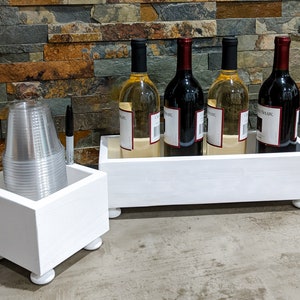 May include: White wooden wine bottle holder with four bottles of wine, two red and two white, and a white wooden cup holder with clear plastic cups.