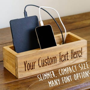 May include: A wooden charging station with a tablet and smartphone inside. The station has the text "Your Custom Text Here!" engraved on the front. The text "SLIMMER, COMPACT SIZE MANY FONT OPTIONS" is also visible.