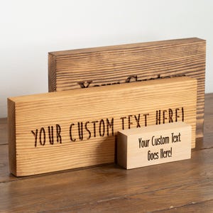 May include: Three rectangular wooden blocks of varying sizes and wood tones. The largest dark brown block has partially visible text. The medium light wood block features the engraved text YOUR CUSTOM TEXT HERE!. The smallest block has printed text.