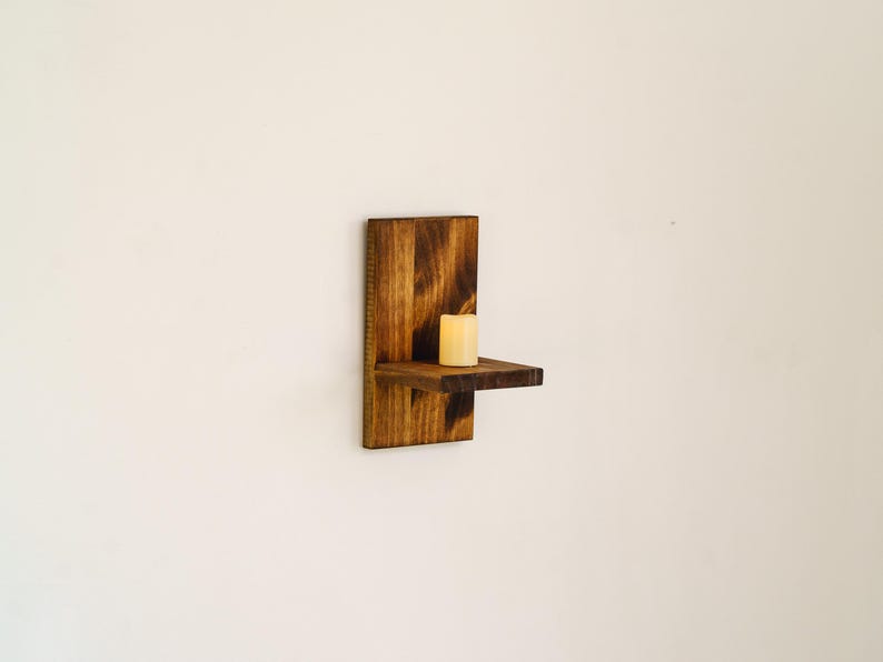 May include: A rustic, wooden wall-mounted candle holder. The holder is stained a warm brown and features a small shelf to hold a single candle. A cream-colored candle sits on the shelf, ready to be lit. The design is simple and elegant.