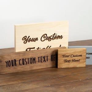 May include: Four rectangular wooden signs of varying sizes and finishes. The largest is light wood with engraved text. A dark brown sign features black text reading YOUR CUSTOM TEXT HERE. A small light brown sign reads Your Custom Text Here!