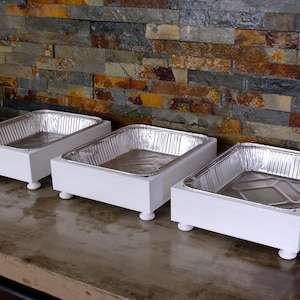 May include: Three white wooden serving trays with aluminum foil pans on top. The trays have four small feet on the bottom and are arranged in a row on a gray surface.