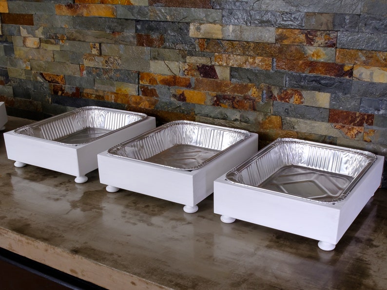 May include: Three white wooden serving trays with aluminum foil pans on top. The trays have four small feet on the bottom and are arranged in a row on a gray surface.