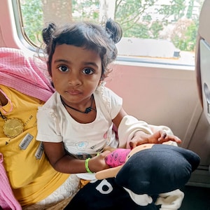 Może przedstawiać: Mała dziewczynka z ciemnymi włosami w kucykach siedzi w pociągu i trzyma czarną i różową pluszową zabawkę. Ma na sobie białą koszulę z kolorowym wzorem i naszyjnik. Dziewczynka patrzy w obiektyw.