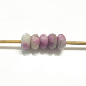 Puede incluir: Cinco cuentas de piedra preciosa rosa y blanca enhebradas en un pincho de madera. Las cuentas son redondas y tienen una superficie facetada.