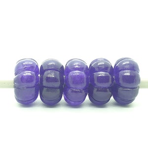 Könnte beinhalten: Fünf violette Glasperlen mit einer strukturierten, unebenen Oberfläche. Die Perlen sind auf einer weißen Schnur aufgefädelt.