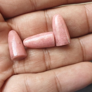 May include: Four pink, cone-shaped stones with a polished finish are displayed in a hand. The stones have a speckled appearance and a rounded base. The color is a light, dusty rose.