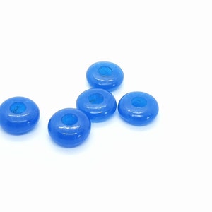 May include: Five translucent blue donut-shaped beads with a hole in the center are scattered on a white background. The beads have a glossy finish and a slightly darker blue center.