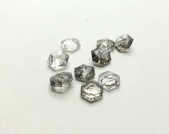 May include: A group of eight clear quartz gemstones with black inclusions. The gemstones are faceted and have a hexagonal shape.