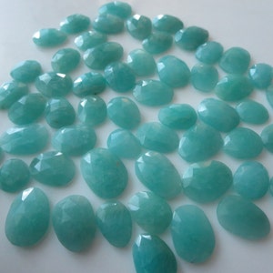 Peut inclure: Plusieurs pierres précieuses d'Amazonite taillées en forme ovale, de tailles et de formes variées. Les pierres sont d'un bleu-vert clair et sont disposées sur une surface blanche. Les pierres ont une surface polie et réfléchissante.