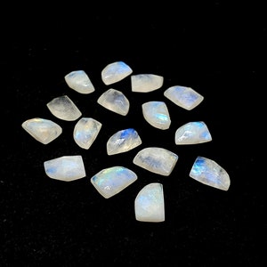 Puede incluir: Una colección de 15 cabujones de piedra lunar blanca iridiscente, cada uno con un patrón natural único. Las piedras son lisas y pulidas, con un brillo sutil.