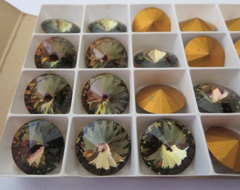 May include: A collection of faceted, iridescent rhinestones in a white divided tray. The rhinestones are various shapes, including round and oval, with a range of colors from gold to olive green. The tray is set against a neutral background.