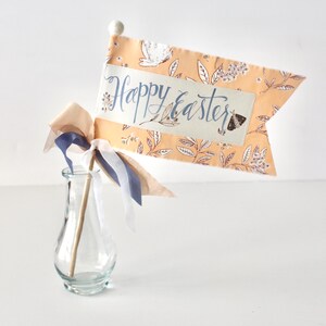 May include: A decorative Easter flag with a peach-colored floral pattern and the words "Happy Easter" in blue script. The flag is attached to a wooden stick and adorned with ribbons in cream, navy, and peach. A small glass vase holds the flag.