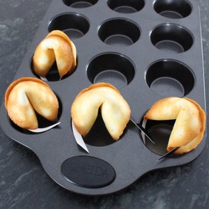 Könnte beinhalten: Eine schwarze Metall-Muffinform mit sechs Glückskeksen darin. Die Kekse haben die Form eines Halbmondes und sind goldbraun.