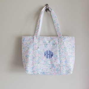 May include: A floral tote bag with a white background and a pattern of small blue and pink flowers. The bag has a front pocket and a blue monogram. The handles are white.