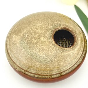 May include: A round, ceramic flower frog with a speckled, tan top and a dark brown base. The frog has a central hole with metal pins for arranging flowers. The design is simple and elegant.