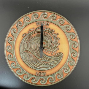 May include: Round, ceramic tide clock with a wave design. The clock face is orange with a green border featuring a repeating swirl pattern. The words "HIGH" and "LOW" are at the top and bottom. The clock hands are black.