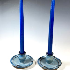 May include: Two ceramic candle holders with blue tapers. The candle holders are light blue with a dark blue rim and a small, round base. The blue candles are tall and slender, adding a pop of color to the scene.