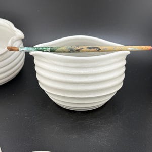 May include: A white ceramic paint bowl with a ribbed exterior, holding a paintbrush. The brush has a wooden handle and a worn appearance. The bowl is part of a set of art supplies.