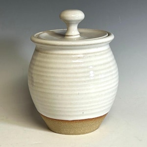 May include: A white ceramic jar with a lid. The jar has a rounded body with horizontal ridges and a tan base. The lid has a small knob on top. The jar is approximately 6 inches tall.