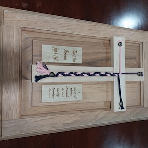 Op de afbeelding: Een houten plaquette met een roze en zwart gevlochten koord bevestigd aan een kruis. De plaquette heeft drie kaartjes met de tekst "For & Fris", "Horseman", "July 14, 1998" en "cord of three strands is not easily broken".