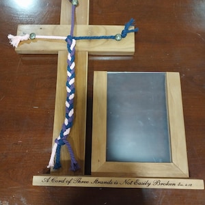 Op de afbeelding: Een houten kruis met een gevlochten touw dat eraan hangt. Het touw is gemaakt van drie strengen roze, paarse en blauwe koord. Het kruis heeft een klein houten plaatje aan de onderkant met de tekst "A Cord of Three Strands is Not Easily Broken Ecc. 4:12".