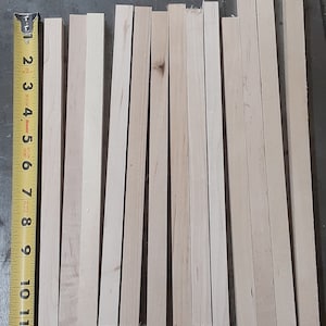 Pode incluir: Um close-up de 12 pinos de madeira, todos com o mesmo comprimento, dispostos sobre uma superfície cinza. Uma fita métrica amarela é visível no lado esquerdo da imagem.