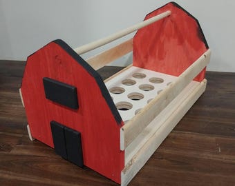 Puede incluir: Una huevera de madera con forma de granero. El granero rojo tiene puertas y ventanas negras, con un interior blanco que contiene agujeros del tamaño de los huevos. Una manija de clavija de madera está en la parte superior y los lados son de madera natural.