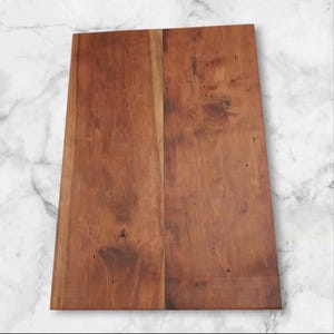 Può includere: Un tagliere rettangolare in legno di colore marrone rossiccio. Il tagliere è composto da due assi unite, che mostrano la venatura naturale del legno e i nodi. La superficie appare liscia e pronta per la preparazione degli alimenti.