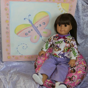 May include: A doll wearing a white and purple pajama top with monkeys and the word "Peace" printed on it. The doll is sitting on a pink and brown beanbag chair with a banana pattern. The doll has brown hair and brown eyes. The doll is sitting in front of a pink and blue picture with a butterfly.