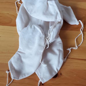 May include: Off-white fabric pouches with drawstring closures. The pouches are made of a soft, textured material and are arranged on a wooden surface. The drawstring cords are white.