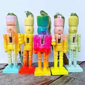 May include: Five colorful wooden nutcrackers in a row, each painted in a different pastel hue. The nutcrackers have a classic design with a soldier's uniform and a decorative ribbon for hanging. The bases are square and painted to match the body.