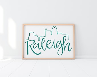 May include: A framed print with a teal blue outline of the Raleigh, North Carolina skyline and the word "Raleigh" written in a teal blue cursive font.