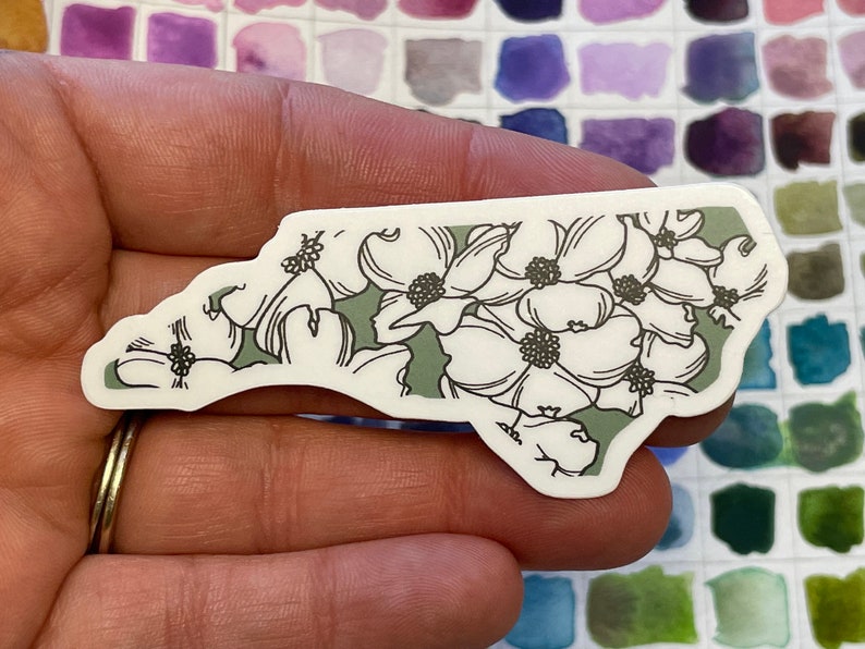 May include: A white sticker with a green outline in the shape of the state of North Carolina. The sticker features a floral design of white dogwood flowers with black centers.