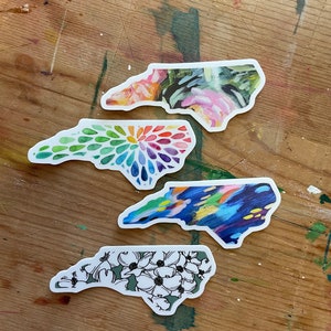 May include: Four North Carolina state outline stickers with different designs. The top sticker has a floral design with pink, green, and white colors. The second sticker has a rainbow design with a circular pattern. The third sticker has a blue and yellow abstract design. The bottom sticker has a green background with white flowers.