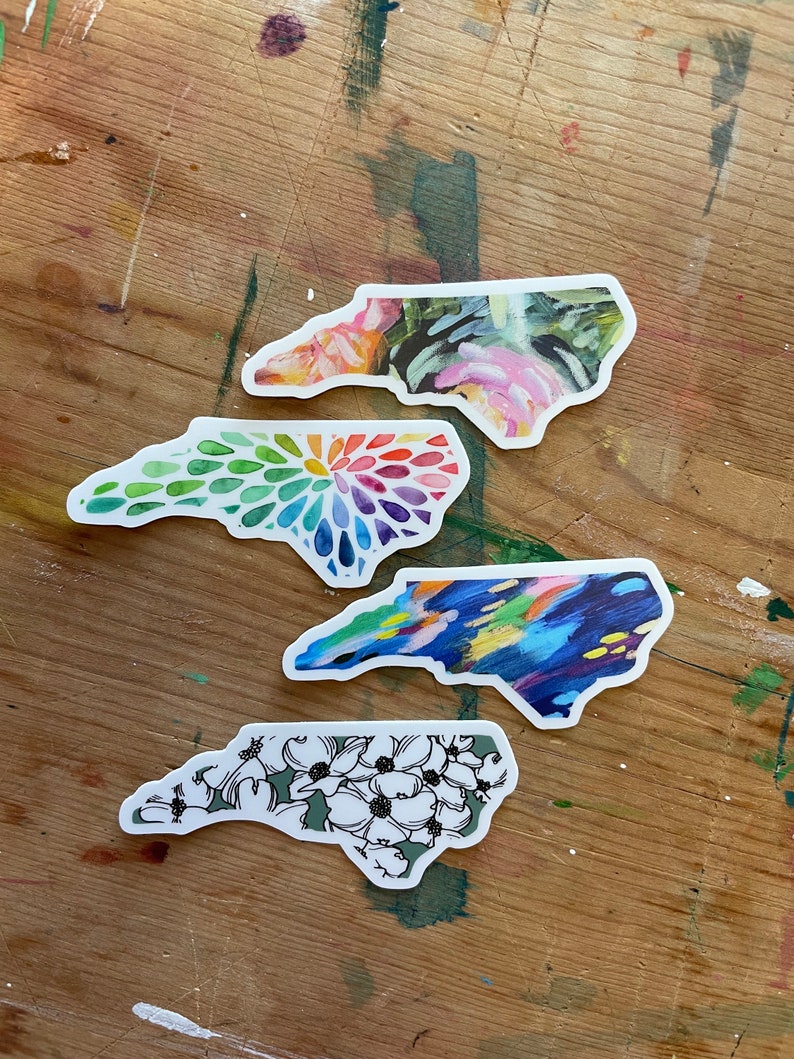 May include: Four North Carolina state outline stickers with different designs. The top sticker has a floral design with pink, green, and white colors. The second sticker has a rainbow design with a circular pattern. The third sticker has a blue and yellow abstract design. The bottom sticker has a green background with white flowers.