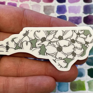 May include: A white sticker shaped like the state of North Carolina with a green background and a white floral pattern.