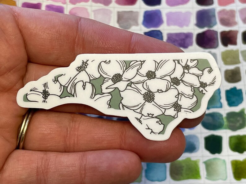 May include: A white sticker shaped like the state of North Carolina with a green background and a white floral pattern.