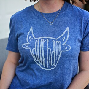 May include: A blue t-shirt with a white graphic of a bull's head with the word "Durham" written inside the head.