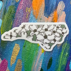 May include: A white sticker with a green outline of the state of North Carolina. The state is filled with white flowers with black outlines and black centers.
