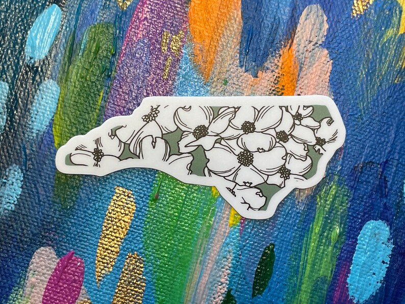 May include: A white sticker with a green outline of the state of North Carolina. The state is filled with white flowers with black outlines and black centers.