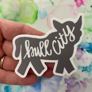May include: A gray vinyl sticker of a bull with the text "bull city" written in white cursive.