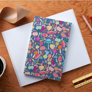 May include: A blue notebook with a colorful mushroom pattern. The mushrooms are in various shades of pink, yellow, green, and orange. The notebook is closed and lying on a white surface.