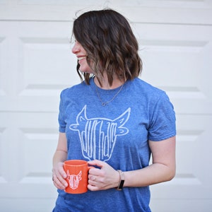 May include: A woman wearing a blue t-shirt with a white graphic of a cow's head. The graphic says "Dur-ham" on it. She is holding an orange mug with the same graphic on it.