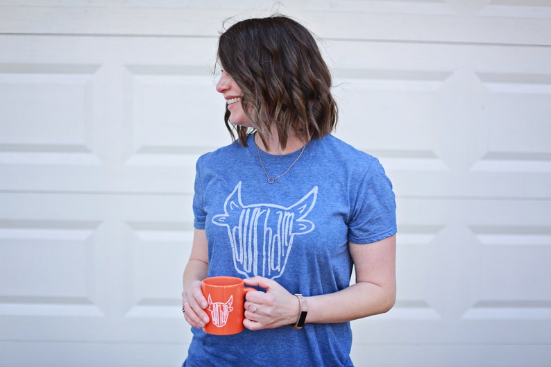 May include: A woman wearing a blue t-shirt with a white graphic of a cow's head. The graphic says "Dur-ham" on it. She is holding an orange mug with the same graphic on it.
