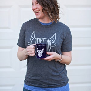 May include: A woman wearing a gray t-shirt with a white bull graphic and blue jeans is holding a dark blue mug with a white bull graphic. The mug has a white handle.