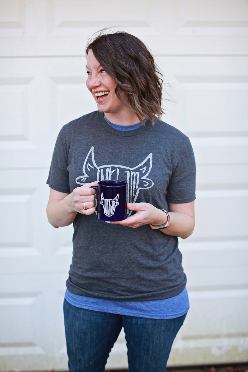 May include: A woman wearing a gray t-shirt with a white bull graphic and blue jeans is holding a dark blue mug with a white bull graphic. The mug has a white handle.